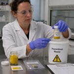A female conservation scientist wearing gloves and goggles places a vial labeled chromate pigment waste into a hazardous waste container in a lab.