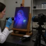 A conservator in a lab uses a UV lamp and camera setup to capture fluorescence on a historic painting to reveal retouching and pigment distribution.